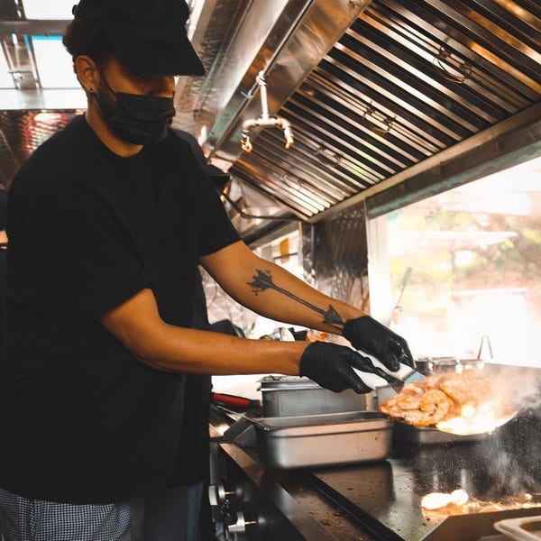 A chef in a black mask and cap cooking hibachi shrimp on a grill with flames.