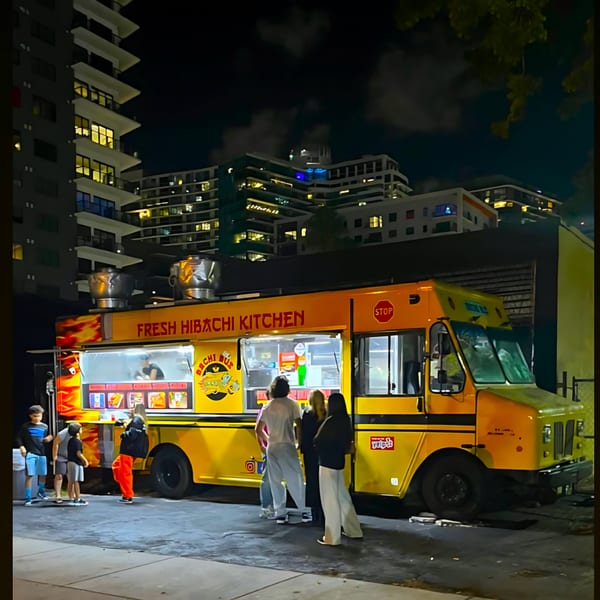 A bright yellow Hibachi Bus Stop food truck at night, with people ordering food.